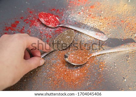 A woman's hand touches a metal spoon with pepper and salt on a gray background with dry spices, on which the sunlight falls. Indian food. Chile. Turmeric. Marjoram. Rosemary. Paprika.