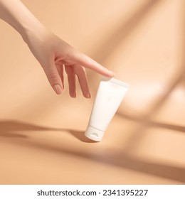 Woman's hand touches a mockup of white tube of cream. Unlabeled packaging for cosmetics in sunlight on beige background. Concept of skincare. Harsh shadows