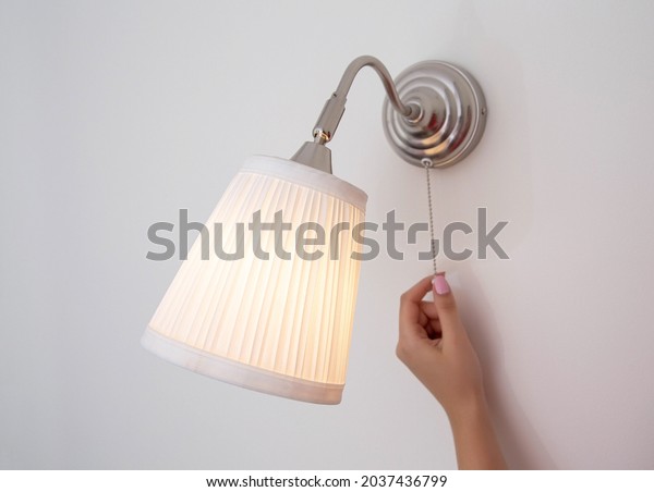 A woman\'s hand turns the wall sconce\
on and off with a rope. Night light above the\
bed