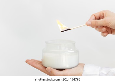 Womans hand uses match to light aromatic candle in glass jar with natural ingredients on white background. Wellness and physical, emotional health concept.