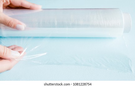 Woman's hand using a roll of transparent polyethylene food film for packing products on the pastel blue table.
