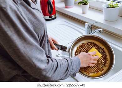 woman's hand washes burnt greasy frying pan with kitchen washcloth in sink. Dirty dishes with burnt food, household chores, washing dishes