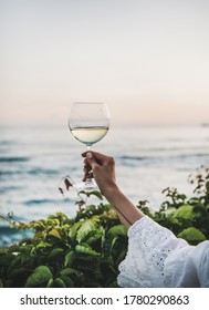 Womans hand in white lace dress sleeve holding glass of white wine with beautiful sunset colors and sea horizon at background. Summer picnic and refreshment drink concept - Shutterstock ID 1780290863