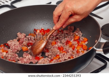 A woman's hand with a wooden spoon stirs raw minced meat with vegetables, which is fried in a black pan.