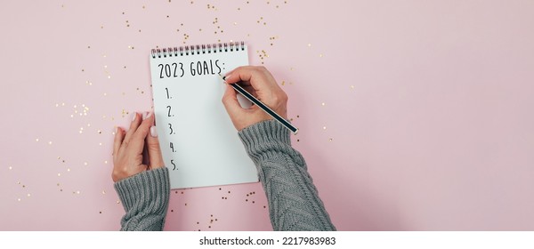 Woman's hand writing 2023 goals in note pad on pink background. Top view, flat lay, copy space. Concept of New Year's plans, goals and actions. Banner image. - Powered by Shutterstock