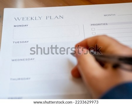 Woman's hand-holding pen and weekly planning on a blank daily schedule. Week planner for organization and goal priorities. Writing down tasks and schedule reminders on office desk.