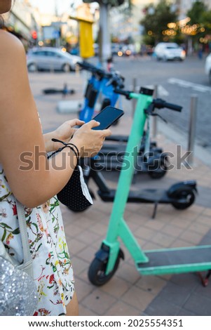 Woman's hands close up holding a phone near the modern city electric kick scooter, going to unlock it. Modern city transportation.