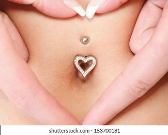 Woman's Hands Forming A Heart Symbol Around Navel With Piercing. Mother Awaiting For The Baby.