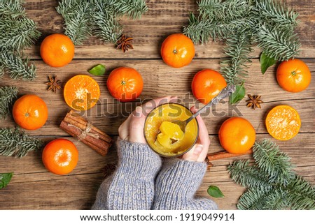 The woman's hands hold sweet jelly made from fresh tangerines. Ripe juicy fruits, aromatic spices, spruce branches. Festive dessert on old wooden boards background, top view