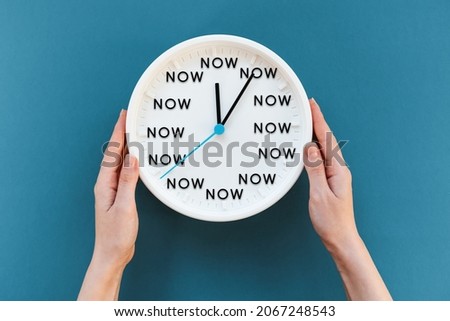 Woman's hands hold a watch with the inscription NOW. Blue background. Flat lay. The concept of time management and procrastination.