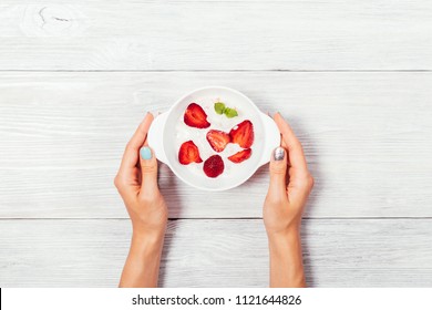 Woman's hands holding a bowl of strawberry ice cream on white wooden table, top view. Cold dessert, decorated with red berries and green leaf mint, simple flat lay composition. - Powered by Shutterstock