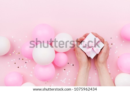 Womans hands holding gift or present box on pink pastel table decorated balloons and confetti. Flat lay style. 