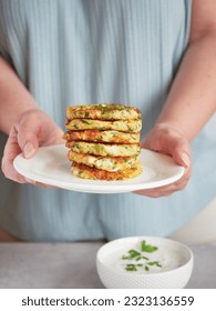 woman's hands holding plate with a stack of vegetable pancakes or fritters, bowl with yogurt and fresh herbs sauce on the table