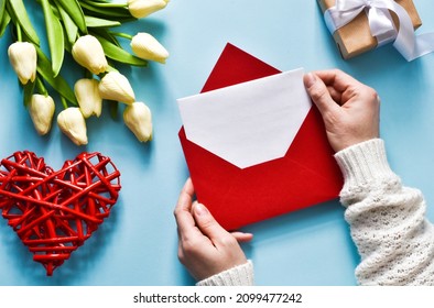 Woman's hands holding red envelope with blank card, white tulips, red heart and gift box. Creative mock up on light blue background. Top view with copy space, flat lay. Greeting card for holiday