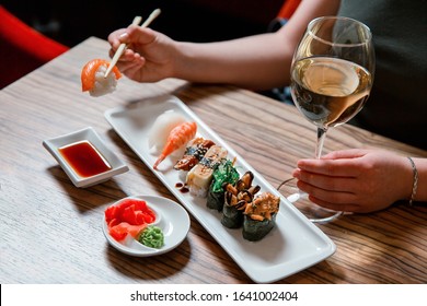 Woman's hands holding sushi with chopsticks and drink white wine, sitting on the wooden table with soy sauce, ginger and wasabi.