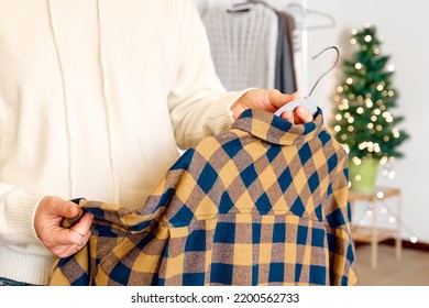 Woman's Hands Holding Warm Plaid Shirt On Hanger On Clothes Rack In The Shop With Blurred Christmas Tree Background. Slow Fashion. Conscious Consumption. Black Friday. Winter Sale.