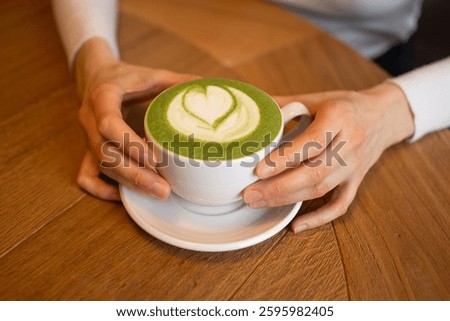 Woman's hands holding a white cup of matcha latte decorated with a heart-shaped latte art, placed on a wooden table, creating a cozy and inviting atmosphere