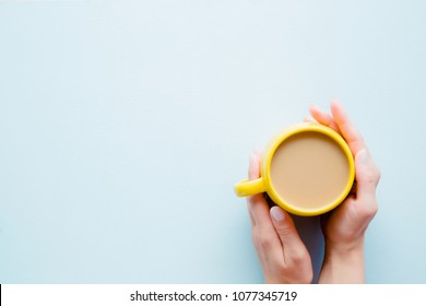 Woman's Hands Holding A Yellow Mug Of Coffee With Milk On Light Pastel Blue Table From Above. Wake Up With Morning Coffee. Empty Place For Inspirational, Motivational Text, Quote Or Logo. Top View.