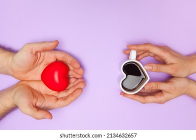 Woman's hands holds heart shaped cup of coffee and man's hands holds red heart. Black hot coffee in a white heart-shaped cup.