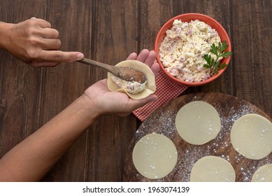 woman's hands making and making capeletis and homemade pasta with circles of dough stuffed with ricotta ham and mozzarella on wooden table and bowl with fresh stuffing