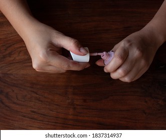 Woman's Hands Opening A Jar Of Pink Nail Polish, With The Lid Removed And The Brush Visible, On A Wooden Table