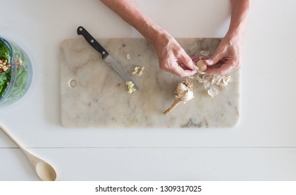 Woman's Hands Peeling Garlic On Marble Chopping Board With Basil Leaves In Food Processor Bowl Nearby (selective Focus)