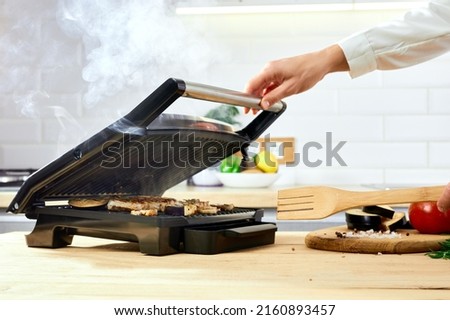 The woman's hands prepares meat on an electric grill on wooden table. Process of cooking meat on an electric grill. Smoke in the home kitchen.