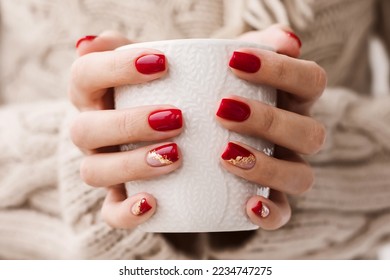 Woman's hands with red manicure and gold foil on the nails. Trendy winter nail design. Woman with a beautiful manicure holding a white knitted cup. The concept of cozy Christmas holidays and New Year.