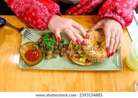 A woman's hands with red nails about to enjoy a bagel sandwich with eggs and potatoes, served with a side of ketchup