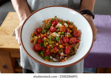 Woman's hands showing a rustic ceramic bowl with quinoa salad with cherry tomatoes, pickles, red and green peppers, cheese, apple, corn, red onion, carrot and chopped cilantro - Powered by Shutterstock