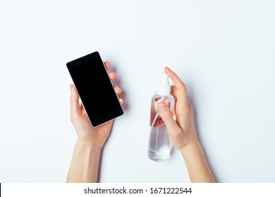 Woman's Hands Spray Disinfectant On Mobile Phone To Clean And Protect Against Viruses, Top View On White Table.