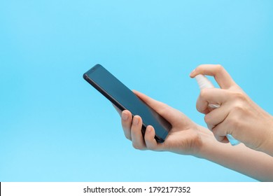 Woman's Hands Spray Disinfectant On Mobile Phone To Clean And Protect Against Viruses. Cleaning Smartphone By Alcohol Hand Sanitizer. Blue Background.