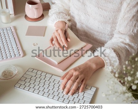 A woman's hands taking notes while using a computer at her desk, a feminine home office scene