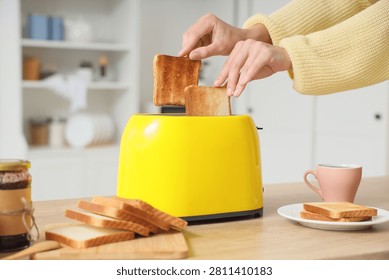Womans hands taking two slices of golden brown toast from a bright yellow toaster on a wooden kitchen counter, the kitchen counter features additional slices of bread on a cutting board, a jar