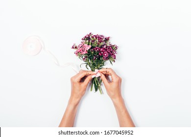 Woman's hands tying satin ribbon bow on small bouquet on white desk, top view.