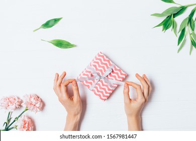 Woman's Hands Unwrapping Small Gift Box Among Fresh Flowers And Leaves On White Wooden Background, Festive Flat Lay.