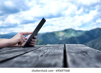 Woman's hands using tablet pc sitting at the old wooden table among the mountains. Green mountains on the background, outdoors, 