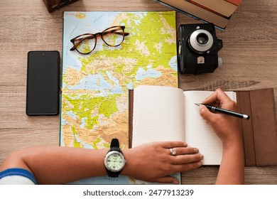 A woman's hands writing in a notebook a list of things needed for vacation, next to a map and a vintage camera - Powered by Shutterstock