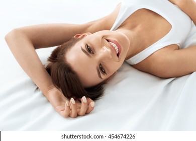 Woman's Health. Closeup Portrait Of Beautiful Smiling Woman With Fresh Face, Soft Skin Having Fun Lying On White Bed. Healthy Happy Girl With Natural Makeup Relaxing Indoors. Beauty, Skin Care Concept