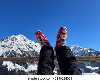 Woman's legs in red ski boots resting on wooden fence after a ski tour on sunny day, snowy mountains in the background. High quality photo