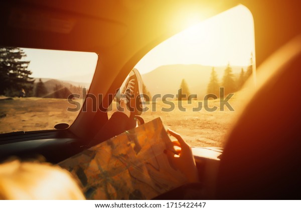 Woman\'s legs in sneakers in the window car with road\
map. Summer travel. Modern hipster girl resting in a car and\
reading a map. Woman with feet on car door. Feet outside the window\
at sunset. 