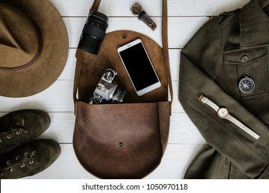 Woman's traveler apparel and  accessories on white wooden background flat lay. Brown leather purse, hiking boots, jawharp, contemporary mobile cellphone, military jacket, watch, film camera, compass.