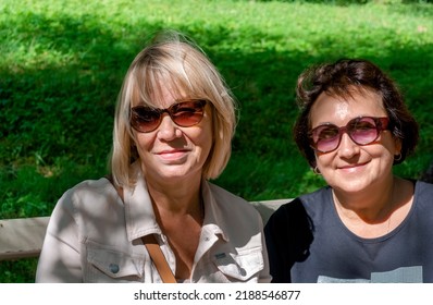 Women 50-60 Years Old In A Good Mood Relax In The Park On A Summer Day. The Sun Illuminates The Faces Of Friends. The Eyes Are Protected From Bright Light By Dark Glasses.