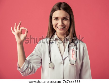 Women against breast cancer. Beautiful female doctor with a pink ribbon on her chest is showing Ok sign, looking at camera and smiling, on red background