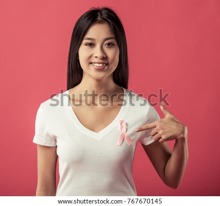 Women against breast cancer. Beautiful Asian woman is pointing at a pink ribbon on her chest and smiling, on red background