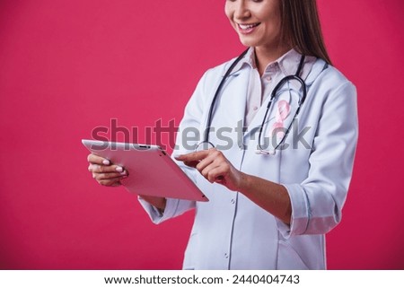 Women against breast cancer. Cropped image of beautiful female doctor with a pink ribbon on her chest is using a tablet and smiling, on red background