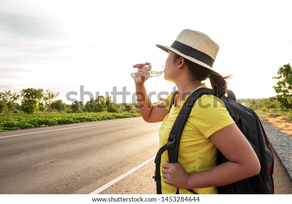 Women backpack tourists, drinking water\
on the highway, with the golden light of the\
sun.