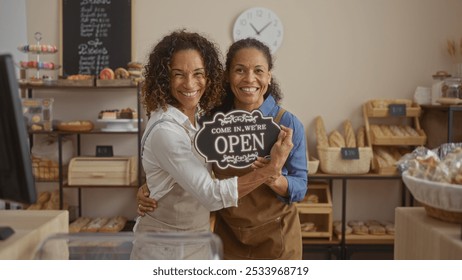 Women bakers smiling in an indoor bakery shop holding an open sign with bread and pastries on display in the background - Powered by Shutterstock