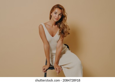 Women beauty. Adorable skinny young female model wears light knitted dress posing seated on chair leaning forward with lovely smile looking at camera, isolated on beige studio background
