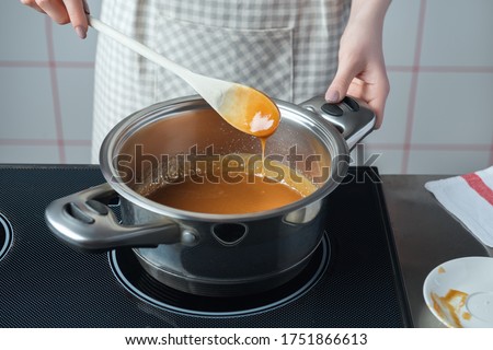 Women in checkered apron with wooden spoon preparing caramel in the stainless steel pan.